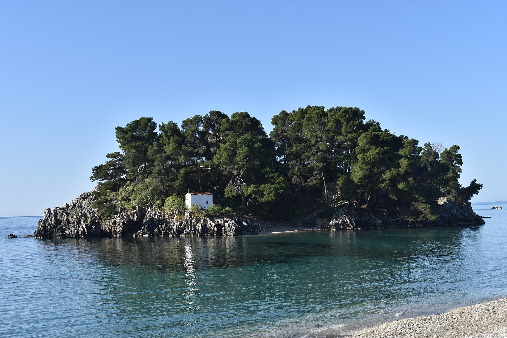 The islet of Panagia at Parga: pines, a small white church, and clear water over a pebble shore.